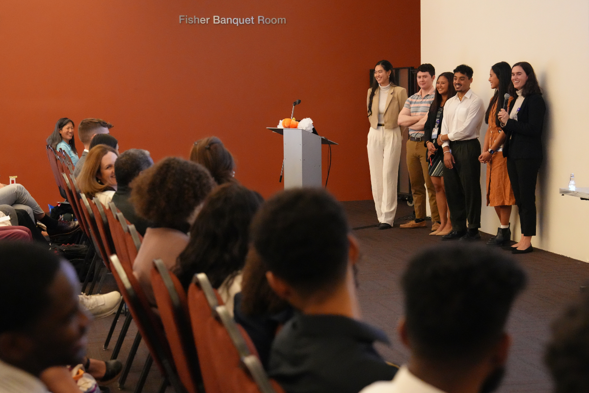 Six students stand next to a podium in front of a seated crowd.