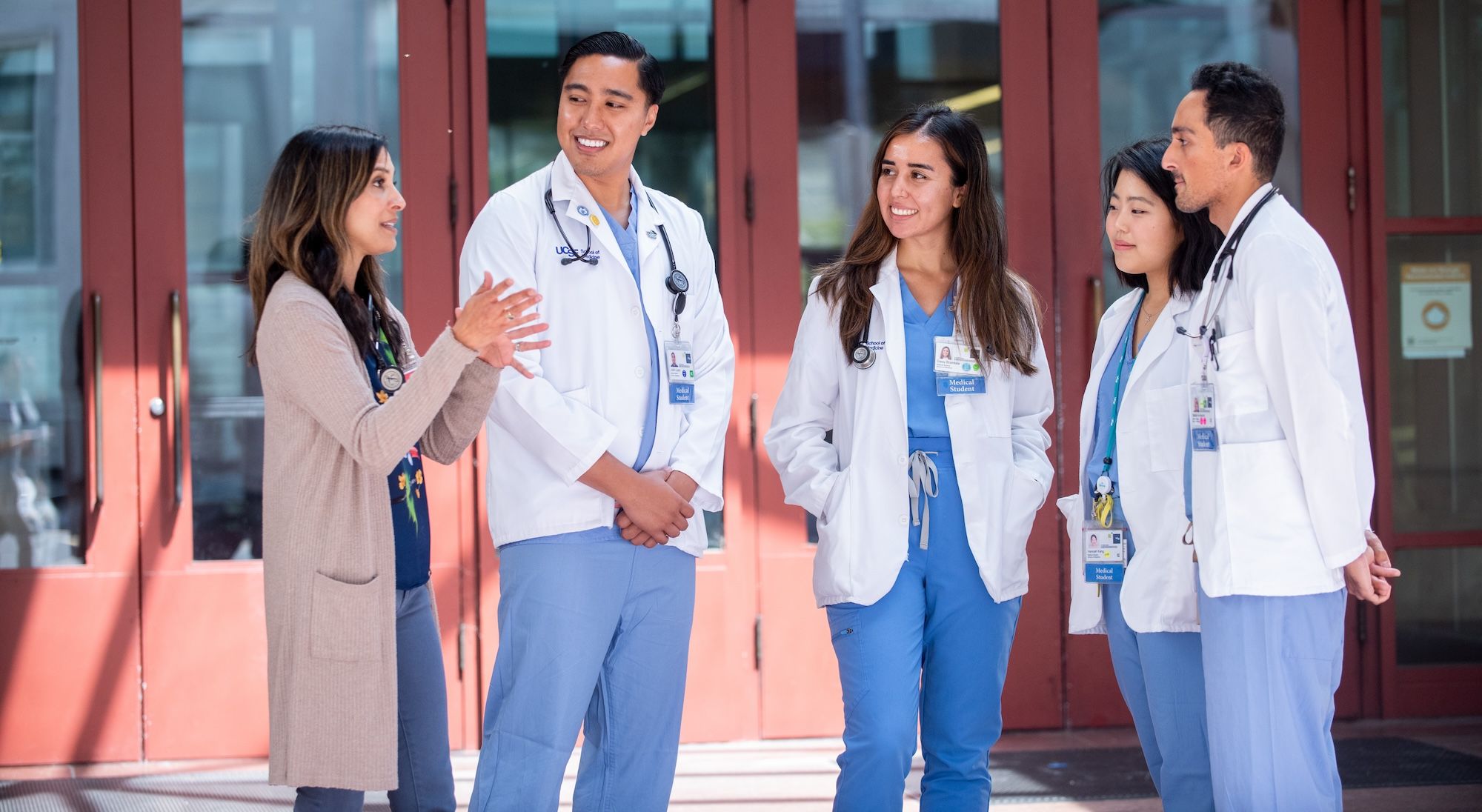 a small group of medical students in white coats listen as an instructor speaks to them in front of the library