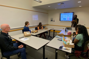 Woman standing next to monitor presents to five people seated at tables surrounding her in a small conference room.