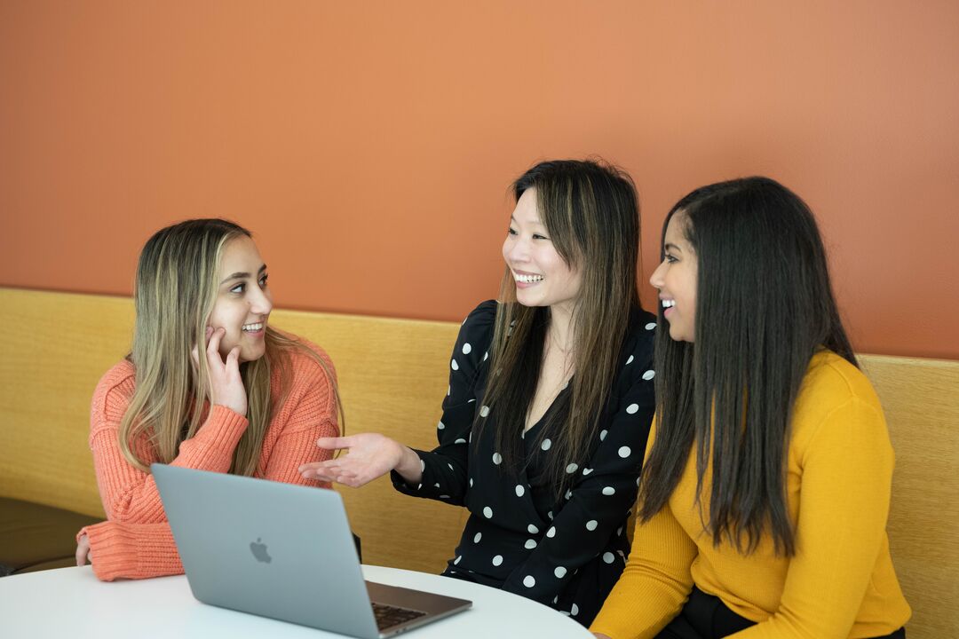 three people smiling and talking around a table with open laptop